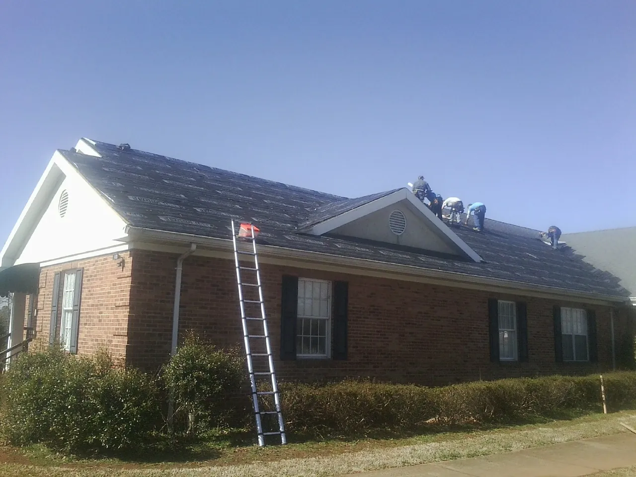 Crew tearing shingles off a brick ranch roof with a ladder set — Metal Roof Repair work by our crew in Hugo